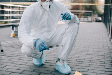 cropped view of male criminologist in protective suit and latex gloves holding tweezers with evidence at crime scene