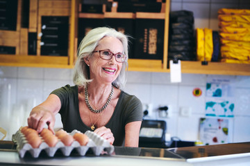 Senior saleswoman smiling while picking brown egg from carton in deli