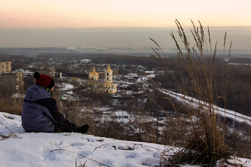 Fototapeta premium In winter, on a frosty evening in the sunset rays of the sun, the boy sits and looks at the city in front of him.