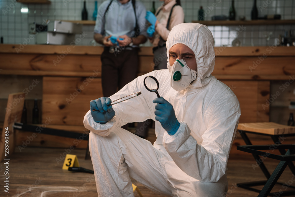 Wall mural focused forensic investigator examining evidence with magnifying glass at crime scene with colleague