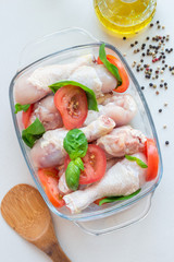 Raw uncooked chicken legs, drumsticks with tomatoes and basil in glass backing dish, meat with ingredients for cooking, vertical, top view