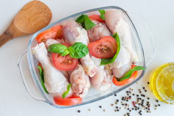 Raw uncooked chicken legs, drumsticks with tomatoes and basil in glass backing dish, meat with ingredients for cooking, horizontal, top view