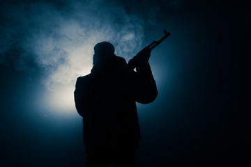 Silhouette of man with assault rifle ready to attack on dark toned foggy background or dangerous bandit in black wearing balaclava and holding gun in hand. Shooting terrorist with weapon theme decor