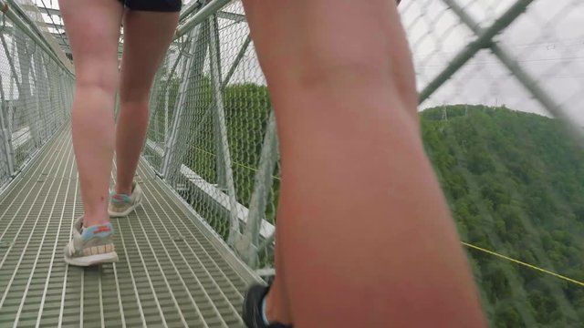 Camera follows caucasian young athletic womans feet in athletic shoes walking on suspended trellis bridge high in mountains against the backdrop of a mountain valley. Close up view