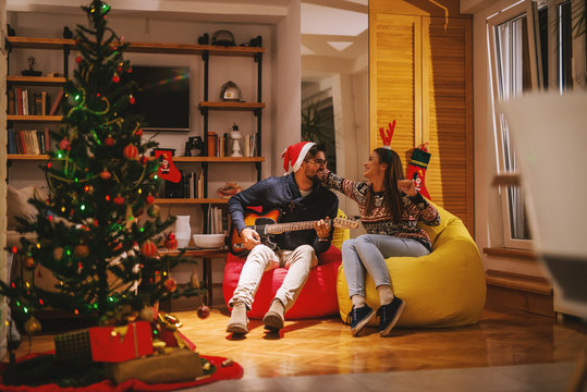 Man playing electric guitar to his girlfriend. On heads reindeer headband and santa's hat. In foreground Christmas tree and presents. Christmas holidays concept.