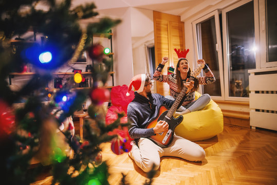 Man playing electric guitar to his girlfriend. On heads reindeer headband and santa's hat. In foreground Christmas tree and presents. Christmas holidays concept.