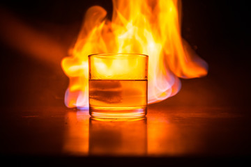Whiskey in fire concept. Glass of whiskey and ice on wooden surface with color light and fog on background. Close up.