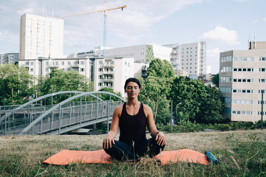 Young woman meditating on field against buildings in city
