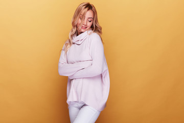 Obraz premium Cute woman with long blond wavy hair dressed in light pink sweater standing on yellow background with her eyes closed