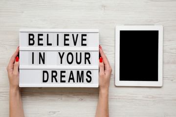 Fototapeta na wymiar Female hands hold modern board with text 'Believe in your dreams', tablet wit blank screen over white wooden background. From above, overhead, flat-lay.