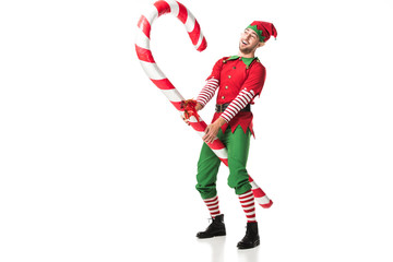 cheerful man in christmas elf costume riding big candy cane isolated on white