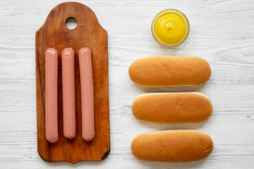 Ingredients for making hotdogs: sausages on wooden board, hot-dog buns and mustard on white wooden table, overhead view. Flat lay, top view, from above.