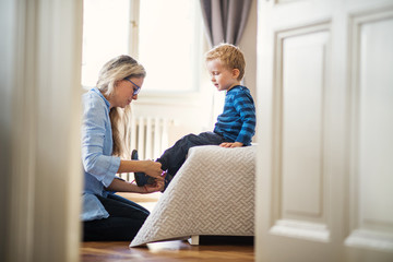 A young mother putting shoes on toddler son inside in a bedroom.