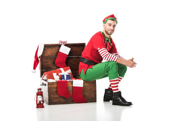 man in christmas elf costume sitting on wooden chest with christmas stockings and presents isolated...