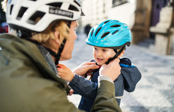 A young father putting on a helmet on his toddler son's head outdoors in city.