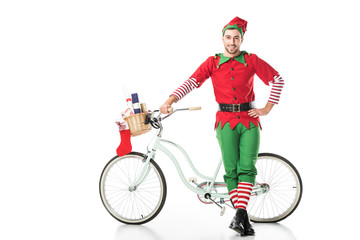 happy smiling man in christmas elf costume with hand on hip standing near bike with basket full of presents isolated on white