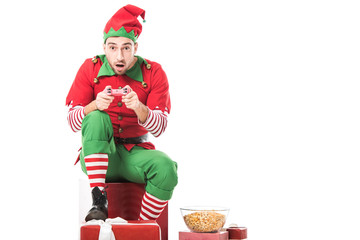 excited man in christmas elf costume sitting on pile of presents and playing video game isolated on...