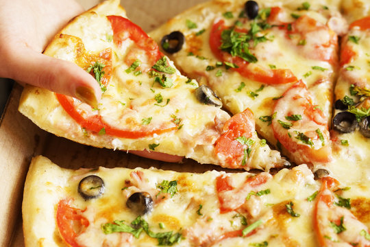 Photo of young woman taking with her hand one slice of hot freshly baked pizza with cheese, ham and chicken, tomatoes and olives out of pizza box, close up   