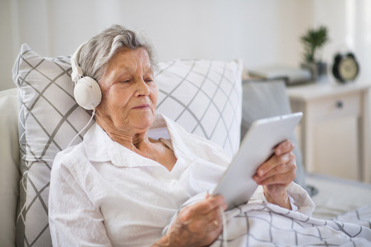 Sick senior woman with headphones and tablet lying in bed at home or in hospital.