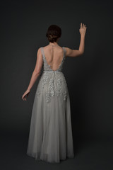 Fototapeta na wymiar full length portrait of brunette girl wearing beautiful long gown, standing pose with background to the camera on grey studio background.