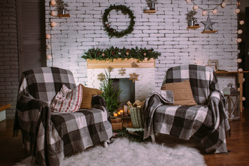 Two armchairs covered with cozy blankets standing near fireplace decorated with garland, candles and basket with gifts. Round green wreath hanging on white brick wall. Christmas festive interior.