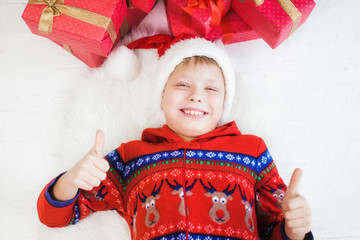 Closeup portrait of cute funny joyful little kid gesturing thumb up as sign of likeness and approval. Photo shoot from above. Kid laying on white wooden floor and fluffy rug. Color horizontal image.