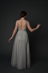 full length portrait of brunette girl wearing beautiful long gown, standing pose with background to the camera on grey studio background.