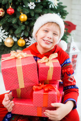 Fototapeta na wymiar Vertical portrait of cute happy smiling european kid with stack of Christmas presents in red wrapping paper. Boy sits near green decorated holiday tree at home. Color photography.