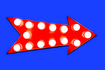 Red arrow shaped vintage colorful illuminated metallic display direction sign with glowing light bulbs isolated on an intense vivid blue background.