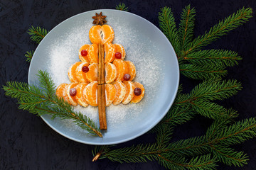 A plate with mandarin slices laid out on it in the shape of a Christmas tree. Black background and spruce branches. View from above. New Year's and Christmas