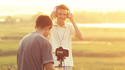 process of photo and video shooting in a field in nature, man making video of guy posing as model in headphones at sunset