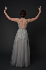 Fototapeta na wymiar full length portrait of brunette girl wearing beautiful long gown, standing pose with background to the camera on grey studio background.