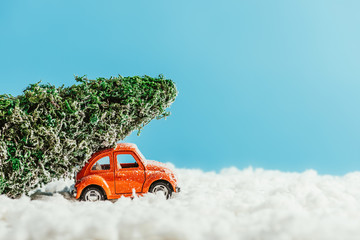 side view of toy car with miniature christmas tree on rooftop riding by snow made of cotton on blue background