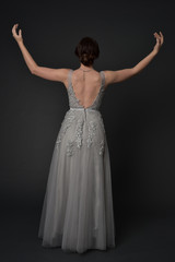 Fototapeta na wymiar full length portrait of brunette girl wearing beautiful long gown, standing pose with background to the camera on grey studio background.