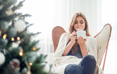 Young relaxed woman resting near Christmas tree and drinking cup of hot tea or coffee