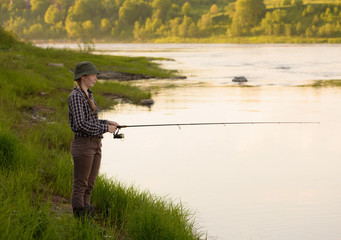 The happy сaucasian girl, 27 years old, is fishing on a riverbank. The fisherwoman is wearing a plaid shirt, the brown trousers, the gumboots and a khaki hat, is holding the spinning in her hands.