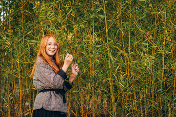 Outdoor close up portrait of beautiful red-haired preteen girl wearing check coat and eyeglasses. Posing in bamboo background