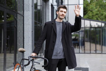 Young business man walking outdoors with bicycle waving.