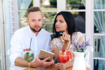 Lovers man and woman sitting at a table in a cafe, socializing drinking cocktails, buying something on the Internet through a tablet. The girl took out her credit card to pay for the online purchase