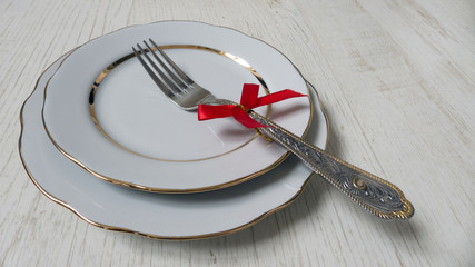 two white plates with a gilded cut edge and a fork with a red bow on a wooden background. festive table setting