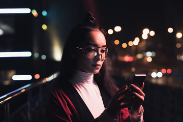 attractive asian girl in burgundy kimono and glasses using smartphone on street with neon light in evening, city of future concept
