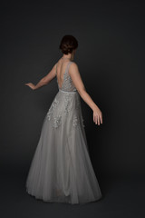 full length portrait of brunette girl wearing beautiful long gown, standing pose with background to the camera on grey studio background.