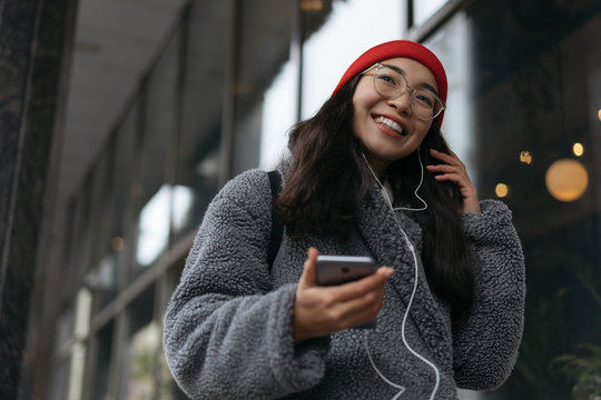 Cheerful happy asian woman in stylish clothing, red hat and hipster eyeglasses listening music outdoors. Portrait of smiling Korean female walking on street, using mobile phone. Positive lifestyle. 