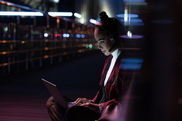 attractive asian girl in kimono sitting and using laptop with headphones on street with neon light,...