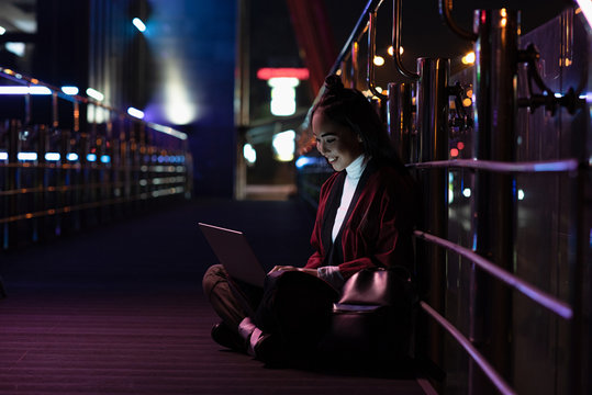 Smiling Attractive Asian Girl In Kimono Sitting And Using Laptop On Street With Neon Light, City Of Future Concept