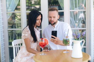 Close-up loving couple sitting at a table in a cafe, a man showing a woman photos on smartphone, a girl interesting look in the phone. Concept of relaxing in a cafe, relationship, people.