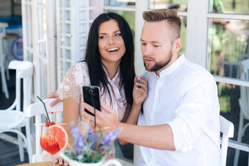 Close-up of beautiful young people sitting in a cafe, taking a photo on the phone. A man makes a selfie with his happy bride. The girl smiles, the man is puzzled. The concept of selfi, gadgets, people
