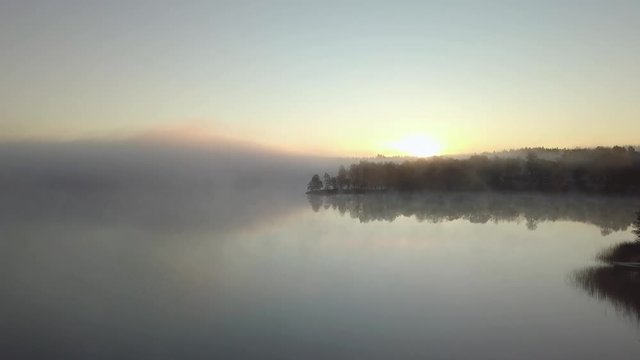 Aerial - Autumn misty morgning in sweden forest. sunrise hitting the trees. big lake in the middle. flying above the fog. 4k