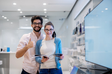 Couple looking st camera ehilr standing in tech store. Man giving thumbs up and woman holding...