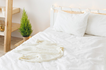 White cozy modern bedroom with holiday decoration. Bed with white bedding set and knitted sweater on it, wooden rack, little Christmas tree in a pot and led garland lights. Home christmas decor.